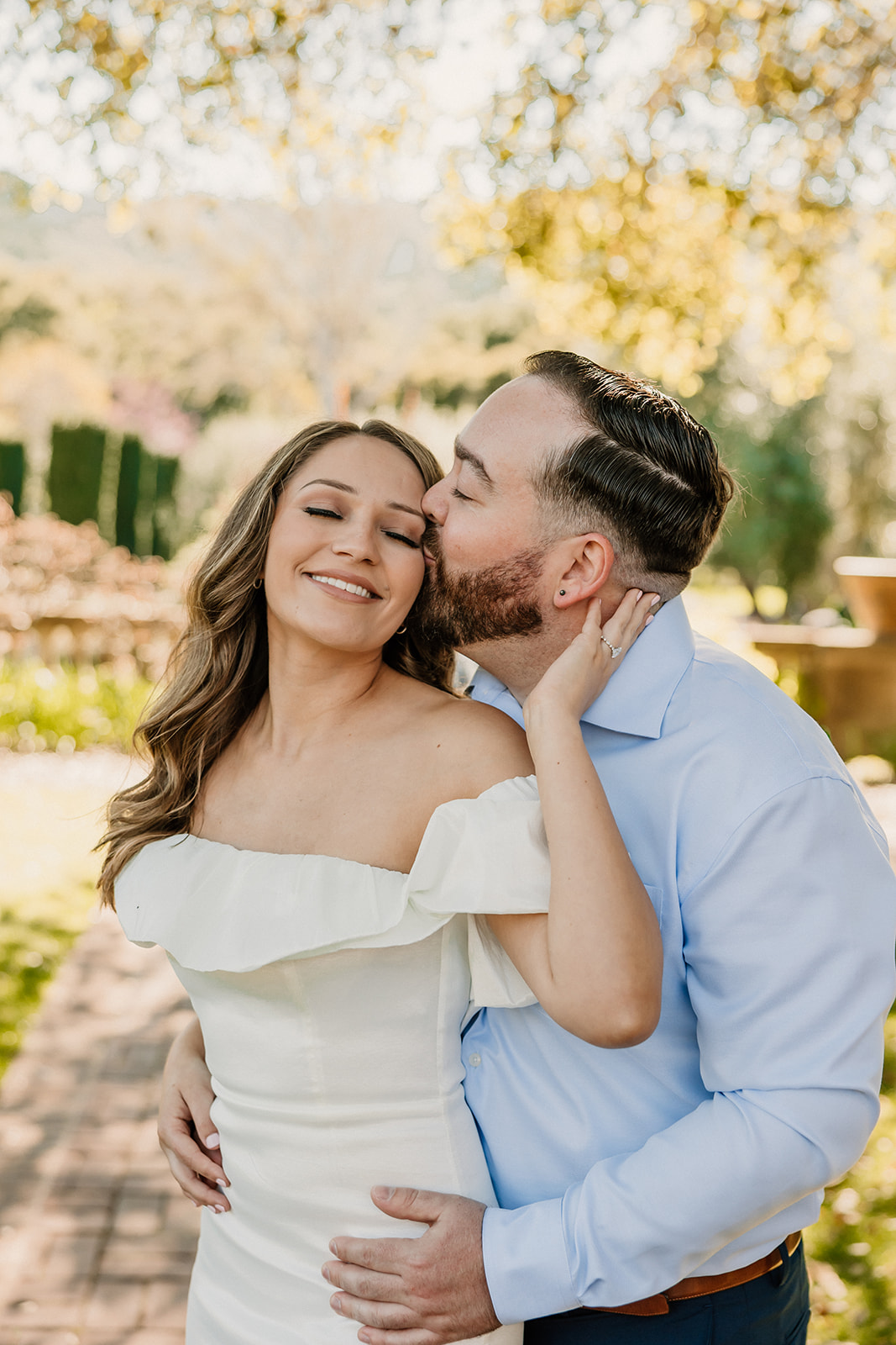 A man in a blue shirt kisses a woman in a white dress on the cheek while standing in a sunlit garden for their engagement photos at the filoli gardens